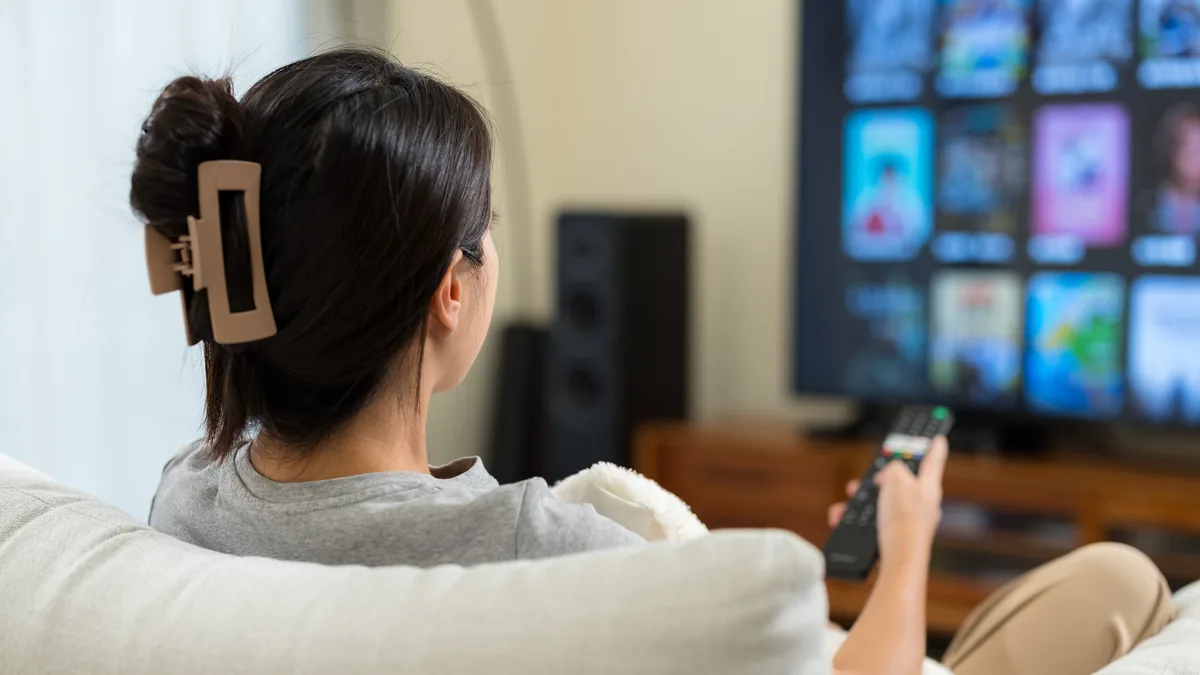 Kvinne i sofaen retter fjernkontrollen mot en TV som viser et rutenett av programbilder