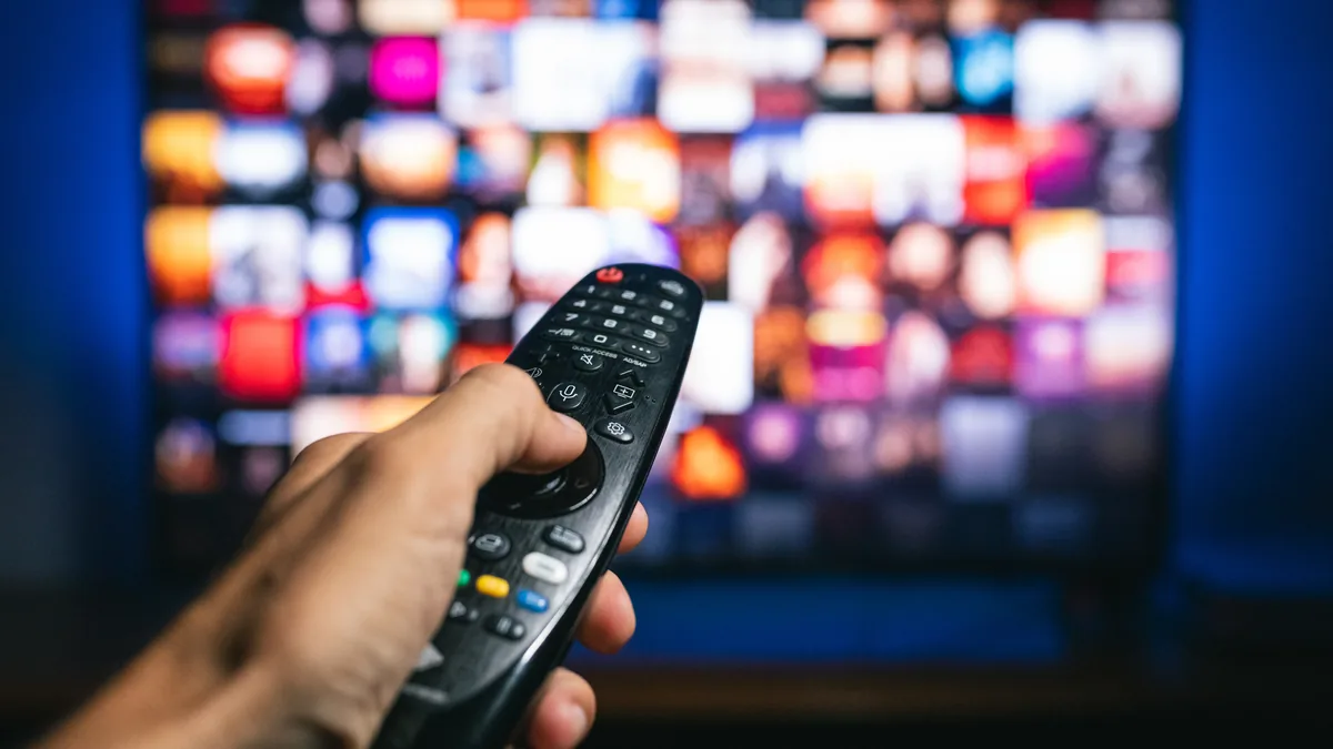 Hånd retter en fjernkontroll mot en TV med et uskarpt rutenett av programbilder