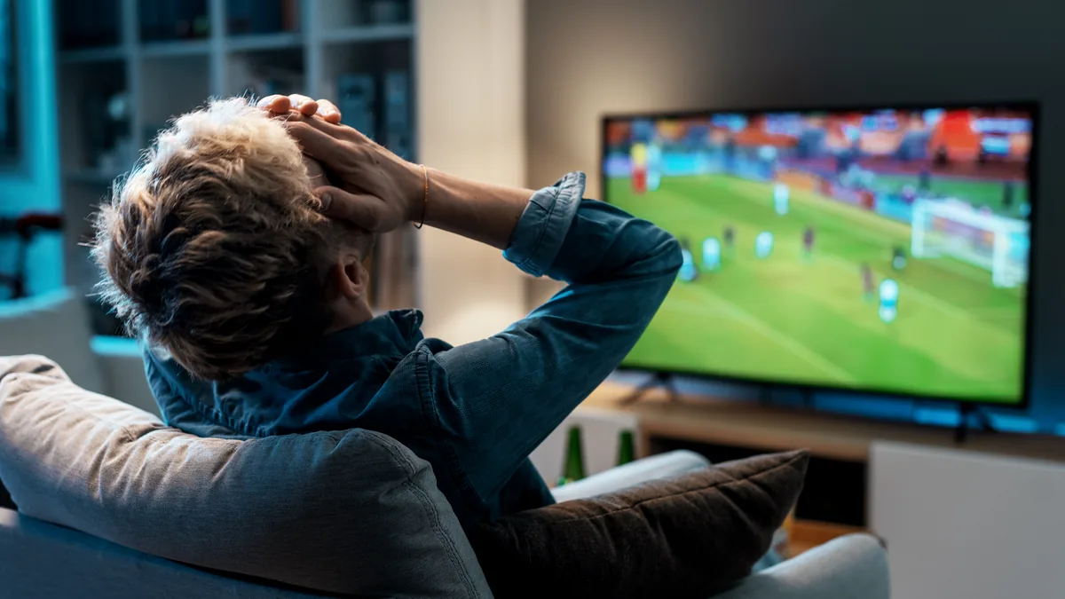 Mann i sofaen holder hendene over hodet mens en fotballkamp går på TV-en