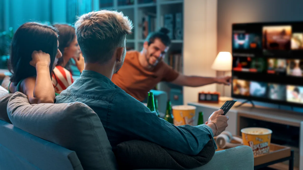 Fire venner i en dempet opplyst stue, én holder fjernkontrollen mot TV-en